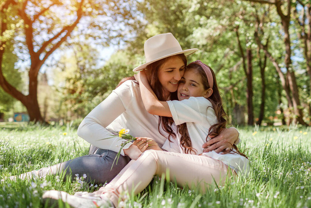 Dzień Matki. Portret matki i jej córki przytulają się siedząc na trawie w wiosennym parku. Rodzinny relaks na świeżym powietrzu zbieranie kwiatów - Zdjęcie, obraz