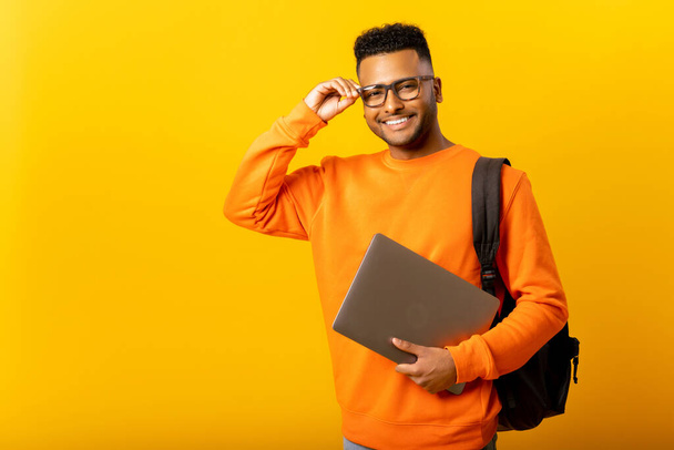 Smart inteligente indian male employee or freelancer man holding laptop and looking at camera with happy smile, isolated on yellow background, arab student with backpack adjusting his glasses - Photo, image