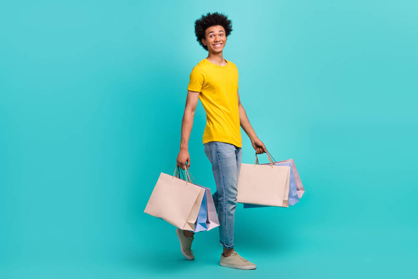 Full body photo of young positive man yellow t-shirt denim jeans hold bags from shopping store advert satisfied isolated on cyan color background. - Foto, immagini