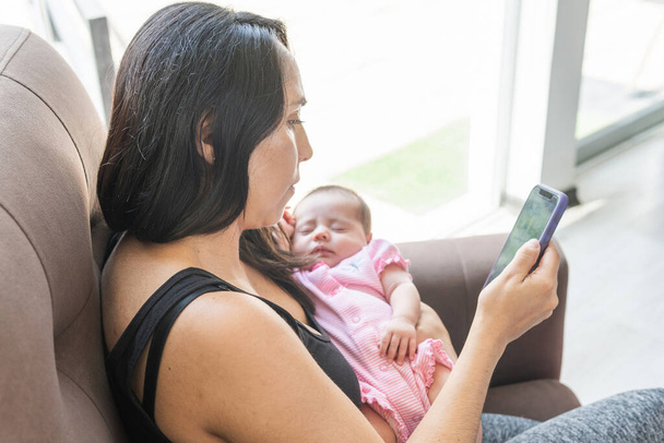 彼女の腕の中で眠っている赤ちゃんと彼女のスマートフォンで作業若い女性 - 写真・画像