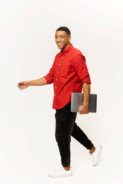 Vertical shot of optimistic handsome man in casual red shirt walking with laptop computer isolated on white background, foreign male student - Fotó, kép