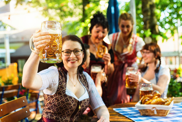 Amigos, dos hombres, tres mujeres, de pie y sentado en el jardín de la cerveza o oktoberfest con vasos de cerveza - Foto, imagen
