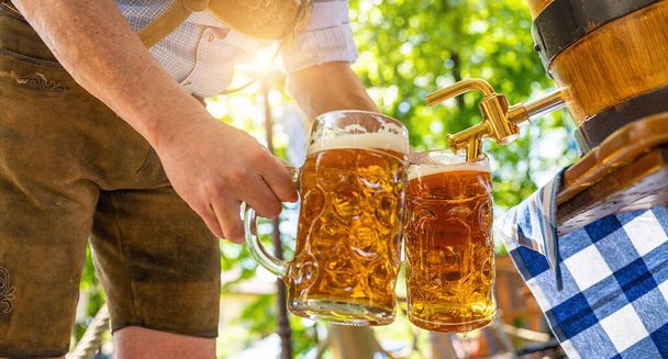 Bayerischer Mann in Lederhosen schüttet im Biergarten aus einem Holzbierfass große Lagerbiere in den Zapfhahn. Hintergrund für Oktoberfest oder Wiesn, Volks- oder Bierfest (für: Ozapft is!)  - Foto, Bild