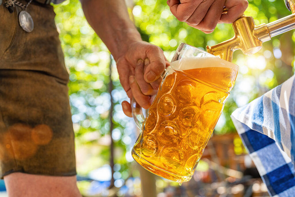 Bavarian man in leather trousers is pouring a large lager beer in tap from wooden beer barrel in the beer garden. Background for Oktoberfest or Wiesn, folk or beer festival (German for: Ozapft is!)  - Photo, Image