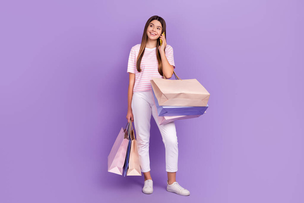 Full size portrait of cheerful teen girl hold bags speak telephone isolated on purple color background. - Photo, image