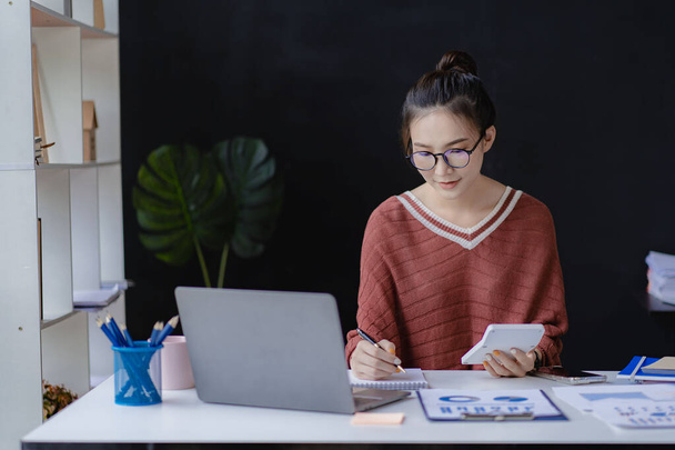 Usmívající se asijská podnikatelka pracující na notebooku u svého stolu v moderní domácí kanceláři dělá výpočty analyzující vykazování výdajů finance - Fotografie, Obrázek