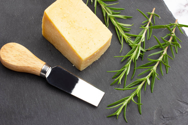 A piece of parmesan cheese, a knife and rosemary twigs on a black slab in a top view - 写真・画像