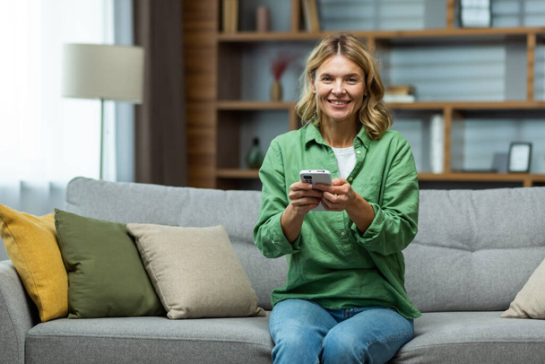 Photo of a senior woman housewife sitting on the sofa and using the phone, smiling at the camera. - Foto, Bild
