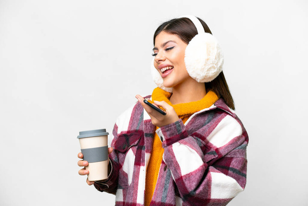 Jonge Russische vrouw dragen winter moffen over geïsoleerde witte achtergrond houden koffie mee te nemen en een mobiele - Foto, afbeelding
