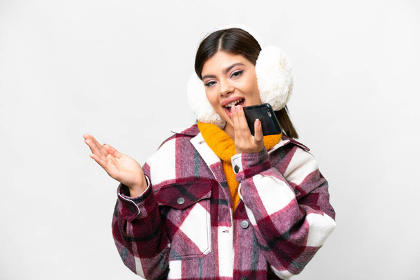 Nuori venäläinen nainen yllään talvi muffinsseja yli eristetty valkoinen tausta pitää keskustelun matkapuhelimen kanssa jonkun - Valokuva, kuva