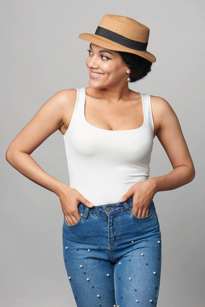 Happy mixed race african american - caucasian woman in jeans and white tank top wearing canotier straw hat with hands in pockets, looking to side smiling - Fotografie, Obrázek