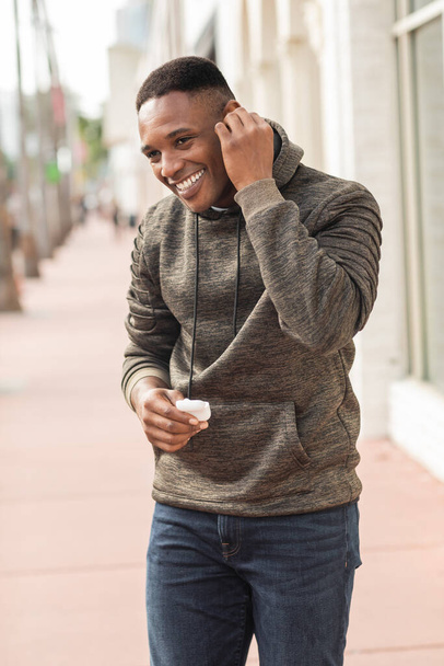 smiling african american man in hoodie holding earphone case on street in Miami  - 写真・画像