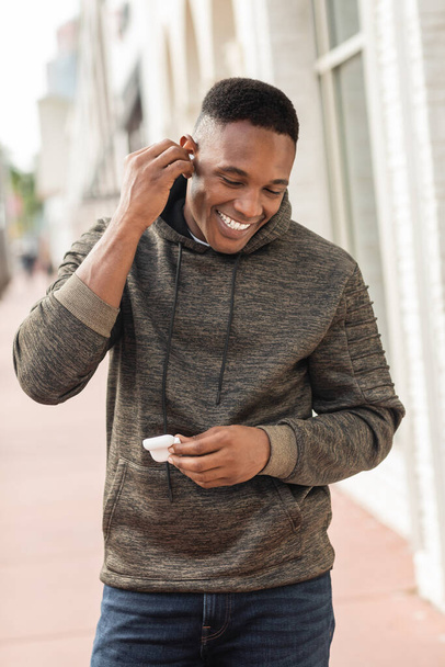 cheerful african american man in hoodie wearing wireless earphone on street in Miami  - Photo, Image