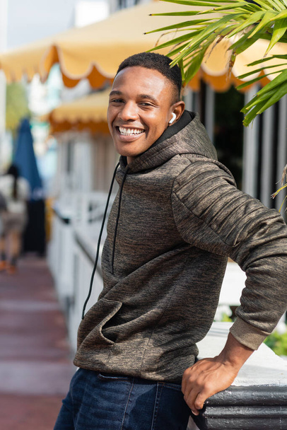 pleased african american man in wireless earphone standing near outdoor bistro  - Foto, imagen