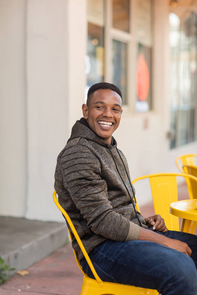 cheerful african american man in hoodie and jeans sitting at bistro table in outdoor cafe  - Фото, изображение