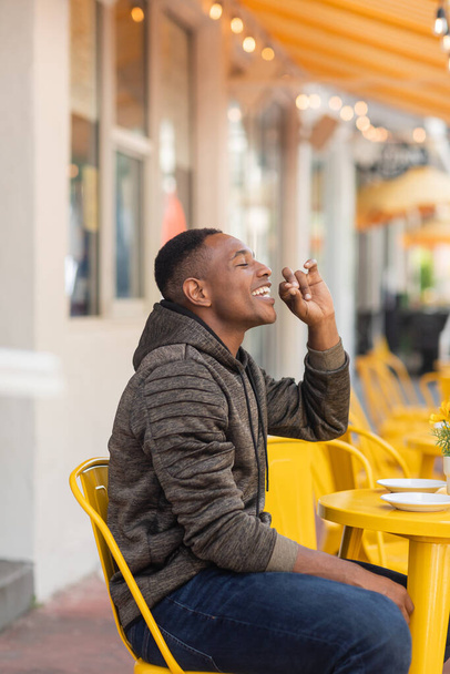 widok z boku zadowolony Afrykański amerykański człowiek w kapturze i dżinsy siedzi przy stole bistro w kawiarni na świeżym powietrzu  - Zdjęcie, obraz