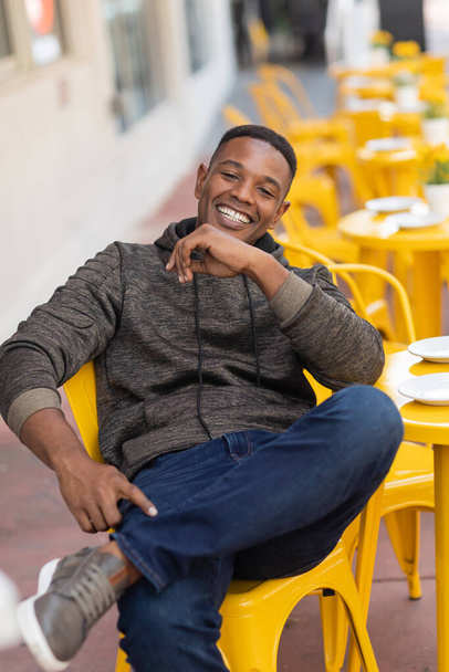 positive african american man in hoodie and jeans sitting at yellow bistro table in outdoor cafe  - Fotografie, Obrázek