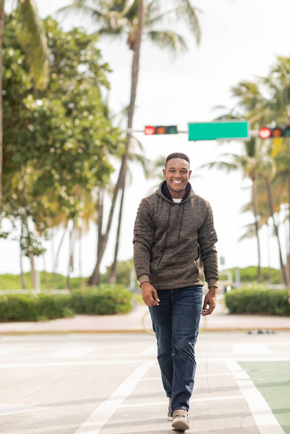 voller Länge von fröhlichen afrikanisch-amerikanischen Mann in Kapuzenpulli und Jeans zu Fuß auf der Straße in Miami  - Foto, Bild