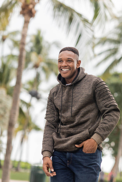 happy african american man in hoodie standing with hand in pocket of jeans - Photo, Image