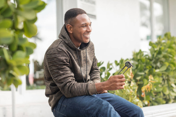 happy african american man in hoodie and jeans holding sunglasses while sitting outdoors  - Foto, afbeelding