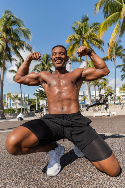 happy african american sportsman in shorts and sneakers showing muscles after workout in Miami beach  - Фото, зображення