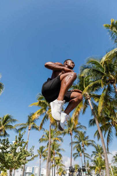 low angle view of shirtless african american sportsman in shorts jumping near palm trees in Miami  - Fotografie, Obrázek