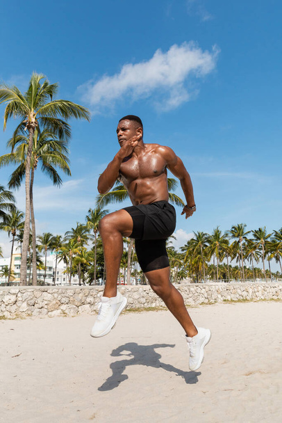 gespierde Afro-Amerikaanse man in korte broek en sneakers rennend op zand in de buurt van palmbomen in Miami strand - Foto, afbeelding