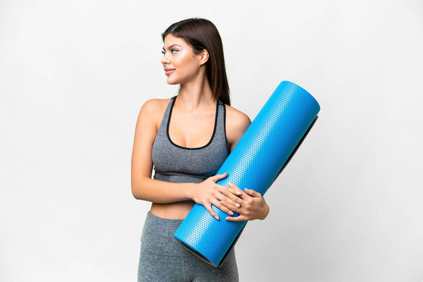 Joven mujer deportiva que va a clases de yoga mientras sostiene una estera sobre un fondo blanco aislado mirando al lado - Foto, imagen