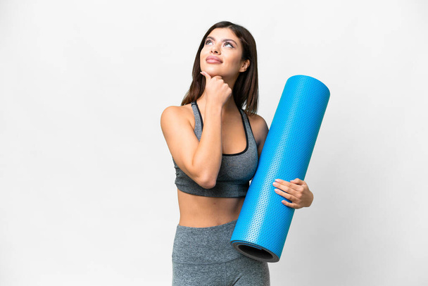 Young sport woman going to yoga classes while holding a mat over isolated white background having doubts while looking up - Photo, image