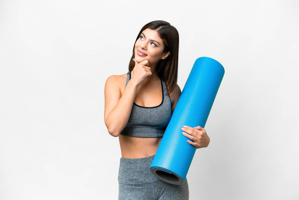 Young sport woman going to yoga classes while holding a mat over isolated white background having doubts - Photo, image