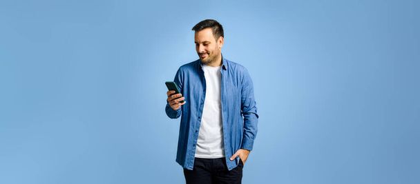 Człowiek z jedną ręką w kieszeni za pomocą smartfona stojąc na niebieskim tle. Przystojny mężczyzna patrząc na urządzenie mobilny ekran. Ma na sobie luźny strój.. - Zdjęcie, obraz