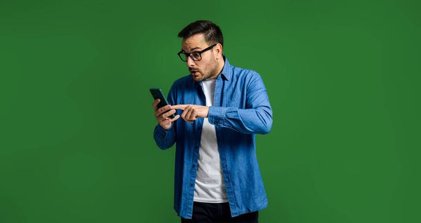 Удивленный человек в джинсах, использующий смартфон на зеленом фоне хромы. Потрясенный парень смотрит на экран устройства, указывая пальцем с широко открытыми глазами.. - Фото, изображение