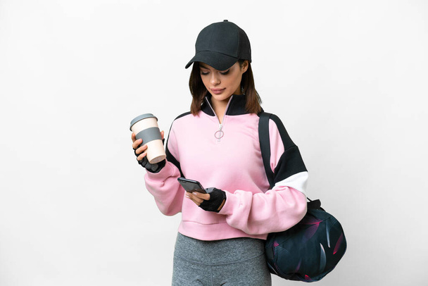 Young sport woman with sport bag over isolated white background holding coffee to take away and a mobile - Φωτογραφία, εικόνα