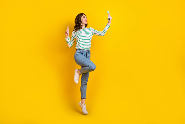 Full length photo of positive girl striped shirt denim pants make selfie on smartphone show v-sign isolated on yellow color background. - Photo, image