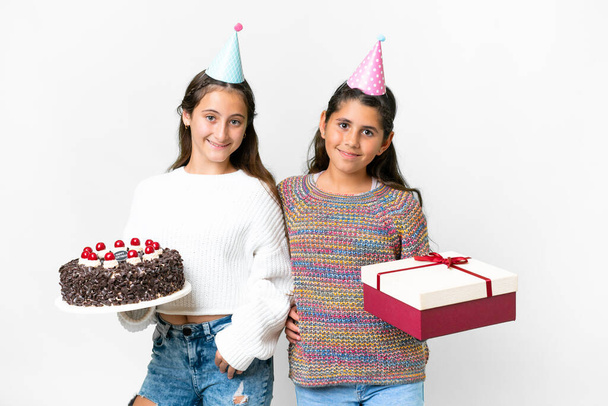 Amigos niñas sosteniendo regalo y un pastel de cumpleaños sobre fondo blanco aislado posando con los brazos en la cadera y sonriendo - Foto, imagen