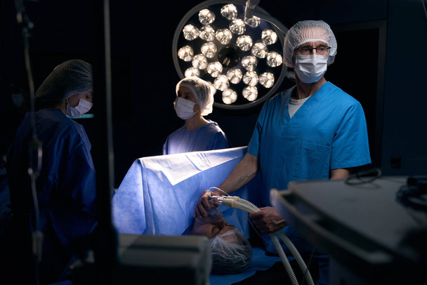 Lekarz anestezjolog trzyma maskę ze znieczuleniem przed twarzą pacjenta, jego koledzy pracują w pobliżu - Zdjęcie, obraz