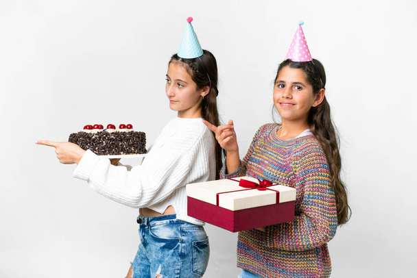 Друзі дівчата тримають подарунок і торт на день народження над ізольованим білим тлом, вказуючи пальцем на бік у боковому положенні
 - Фото, зображення