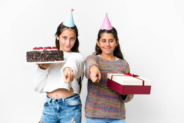 Amigos niñas sosteniendo regalo y un pastel de cumpleaños sobre aislado fondo blanco señala el dedo en usted con una expresión de confianza - Foto, imagen