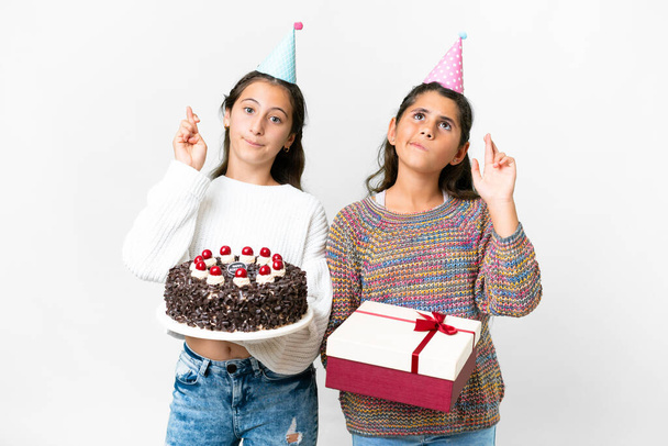 Friends girls holding gift and a birthday cake over isolated white background with fingers crossing and wishing the best - Foto, afbeelding