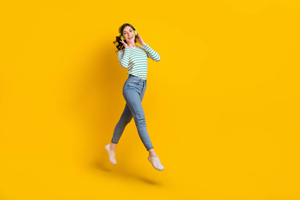 Full length photo of good mood nice girl wear striped shirt denim pants flying touching headphones isolated on yellow color background. - Fotó, kép