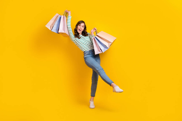 Full length photo of optimistic impressed girl wavy hairdo striped shirt raising up shopping bags isolated on yellow color background. - Photo, Image