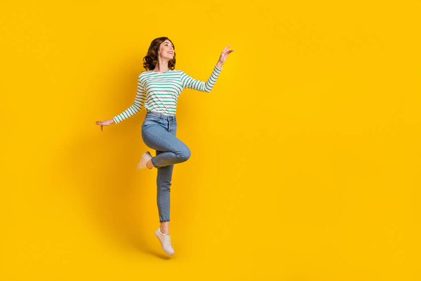 Full size photo of friendly gorgeous woman dressed striped shirt jeans shoes flying look empty space isolated on yellow color background. - Zdjęcie, obraz