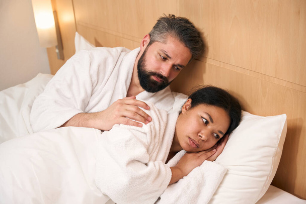 Serious young male lying in bed while touching shoulder of his sad female companion - Photo, image