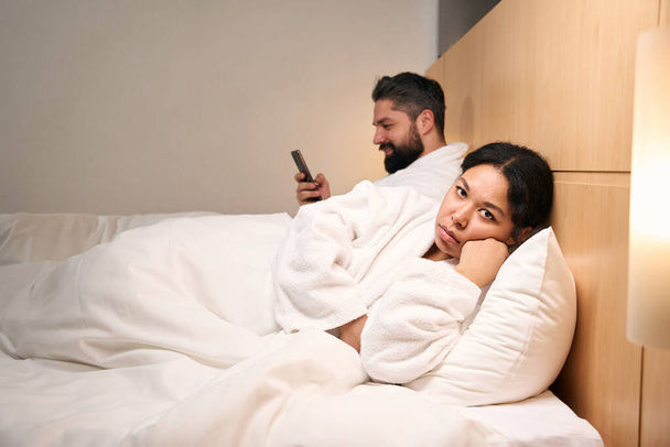 Depressed female lying in bed beside her pleased companion with mobile phone - Valokuva, kuva