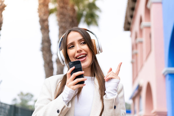 Young pretty woman at outdoors listening music with a mobile and singing - Foto, imagen