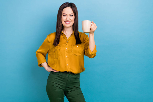 Φωτογραφία της ώριμης επιχειρηματία κυρία φορούν κίτρινο πουκάμισο πράσινο παντελόνι κρατήσει κούπα φρέσκο καφέ ποτό Καλημέρα ζητωκραυγές απομονώνονται σε μπλε φόντο χρώμα. - Φωτογραφία, εικόνα