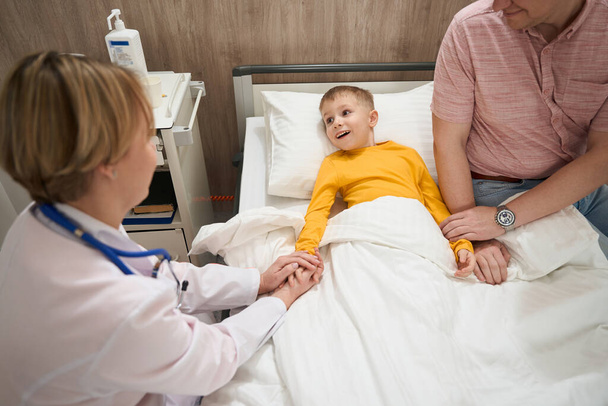 El retrato de vista frontal del niño sonriente en suéter amarillo se comunica con la doctora en la habitación del enfermo. - Foto, imagen