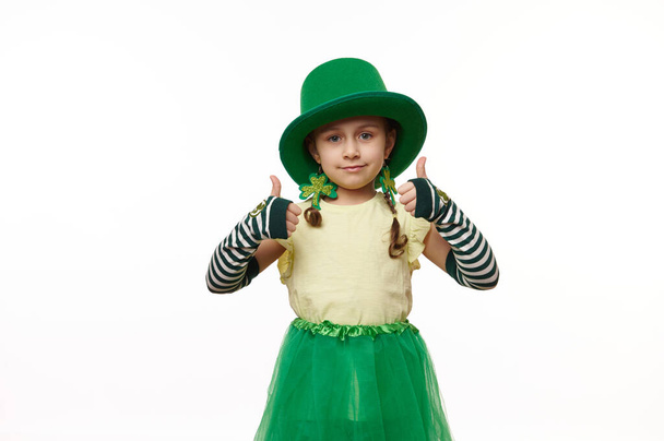 素敵な小さなレプラコーンの女の子カーニバルの衣装に身を包んだ、手袋、緑の帽子、スカートやクローバーの葉のイヤリングを剥ぎ取り、カメラを見て親指を、孤立した白い背景に。聖パトリックの日 - 写真・画像