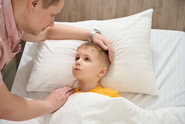 close-up portret van depressieve kleine jongen ligt op medisch bed terwijl op zoek naar zijn vader in de ziekenzaal - Foto, afbeelding