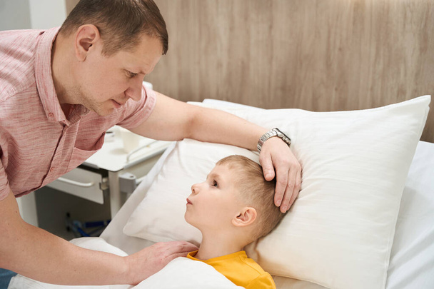 Πλάγια άποψη πορτρέτο του πανέμορφο νευρικό μικρό αγόρι μιλάει με τον πατέρα του στην ιατρική πτέρυγα - Φωτογραφία, εικόνα